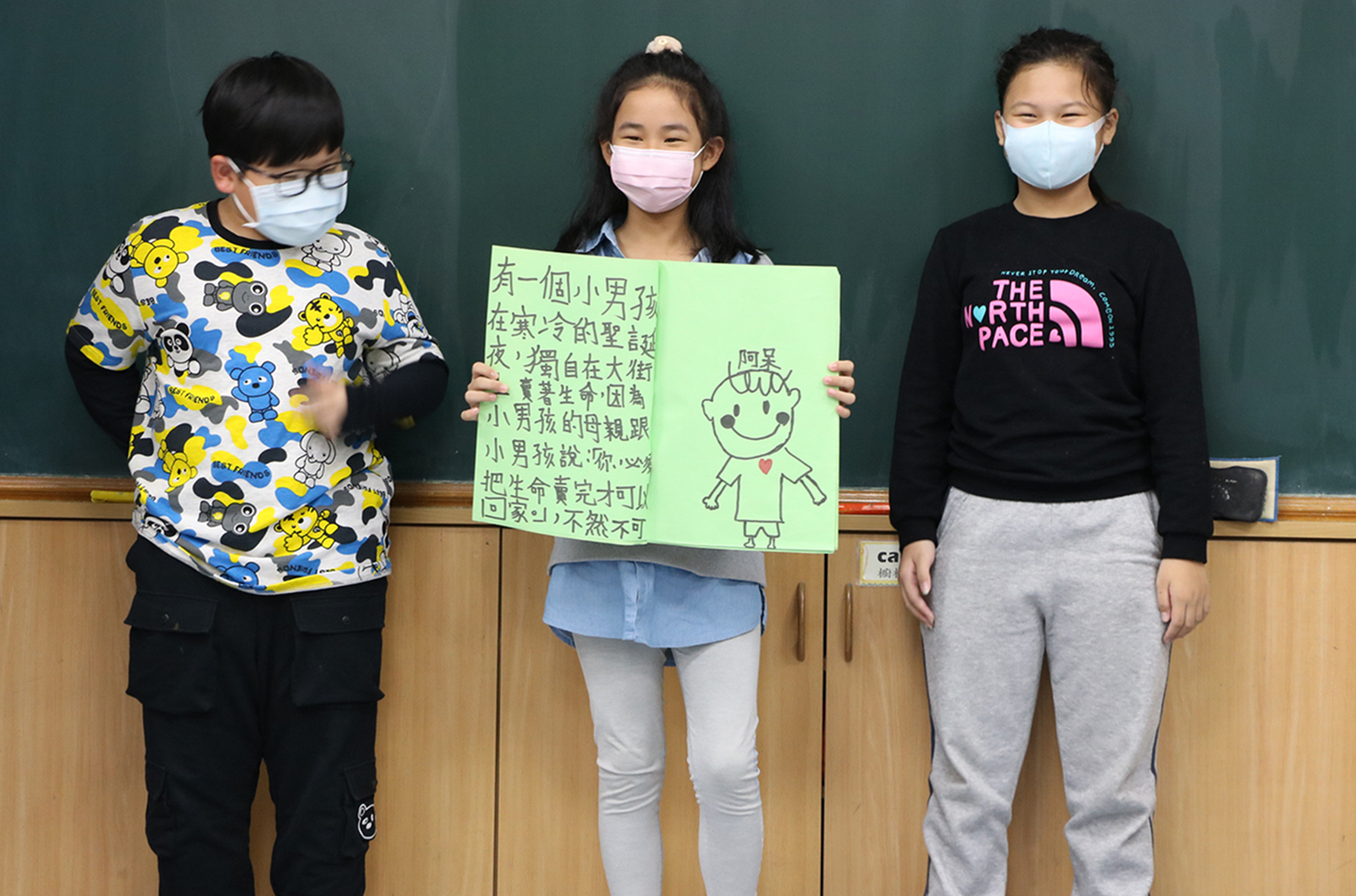 三名戴口罩的學童在教室前合照。中間女童舉著綠色紙張，上面用中文寫著故事內容，並繪有笑臉人物圖。左側男童穿著寶可夢圖案上衣，右側女童穿著黑色北面上衣。背景為綠色黑板。