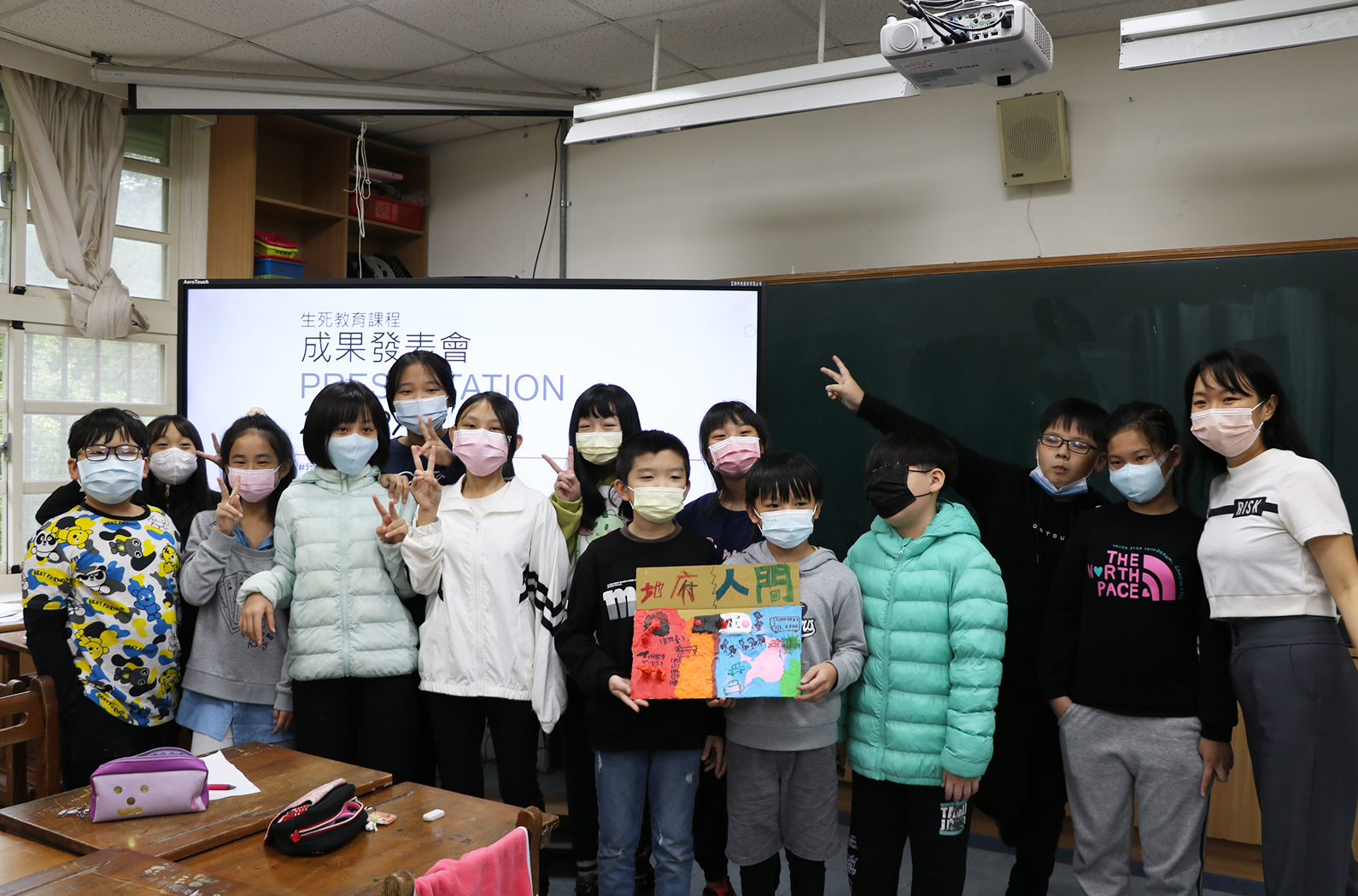 教室內約十五名學生戴著口罩，在講台前排成一列合照。背景投影幕上顯示「生死教育課程成果發表會」字樣。中央學生舉著彩色手工藝作品。教室內有黑板、講台和投影機等教學設備。