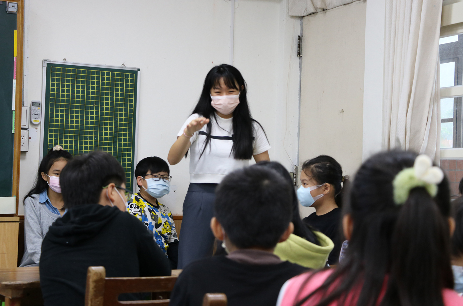 教室內，一位戴著口罩的女性教師站在講台前，面向坐著的學生們進行教學。教室背景有綠色網格黑板，學生們穿著冬季制服，多數戴著口罩。這是一堂師生互動的課堂教學場景。