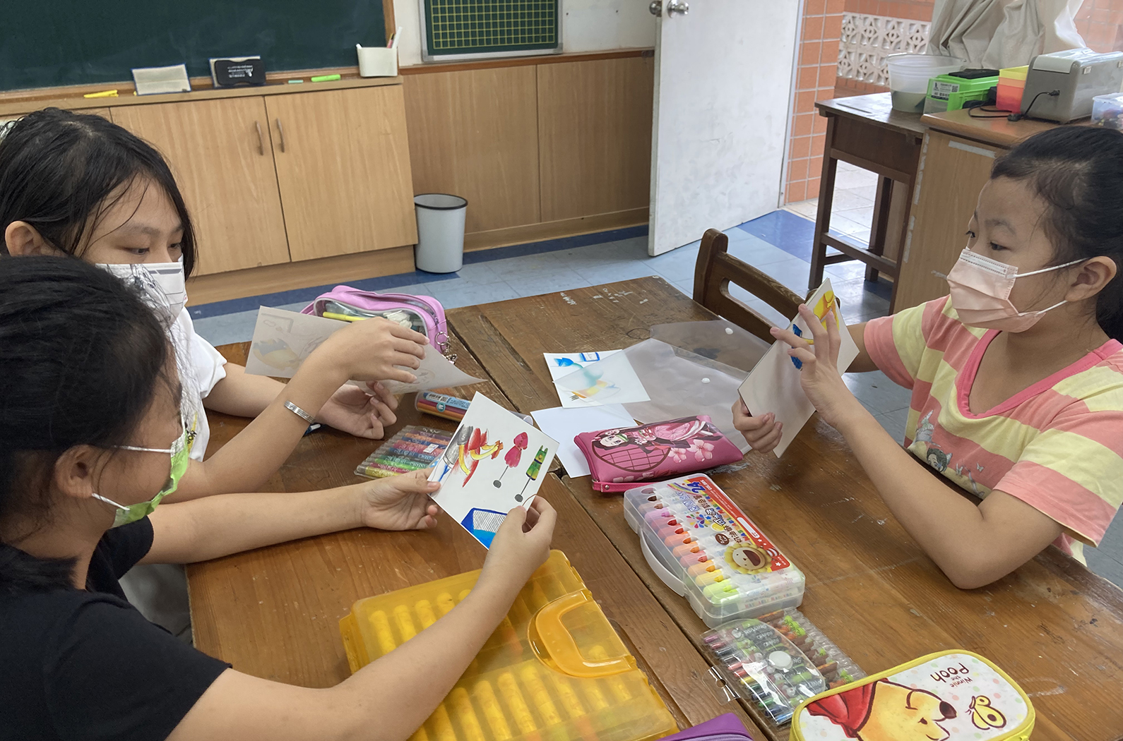 教室內，兩名戴著口罩的小學生坐在木桌前進行美術課。左邊學生穿著黑色上衣，右邊學生穿著粉紅色條紋衣服。桌上放置彩色筆、畫紙、粉紅色背包和美術用具。兩人正專注於繪畫和手工藝活動。