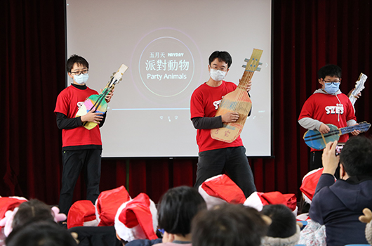三名穿著紅色背心的表演者在舞台上展示樂器，背景投影螢幕顯示「派對動物」主題文字。台下觀眾戴著紅色帽子觀看表演，舞台背景為紅色布幕。
