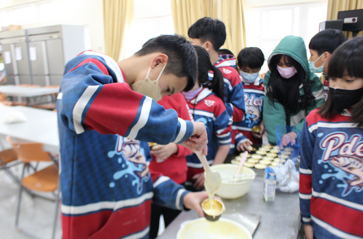 一群穿著紅藍相間運動服的學童在室內聚集，正在進行烹飪或食物製作活動。他們圍著桌子，有人正在處理食材或倒入液體，戴著口罩，認真參與這項集體手工藝活動。
