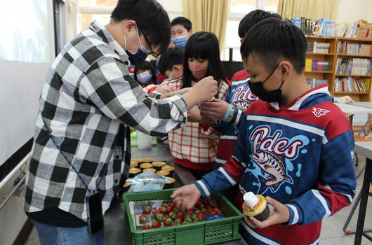 在圖書館內，一位穿著格紋襯衫的成人與多名學生進行食品相關活動。學生們穿著運動服，聚集在綠色塑膠籃周圍，籃中裝有蔬菜和食材。眾人正在挑選或檢視食物，進行互動式的教學或體驗活動。