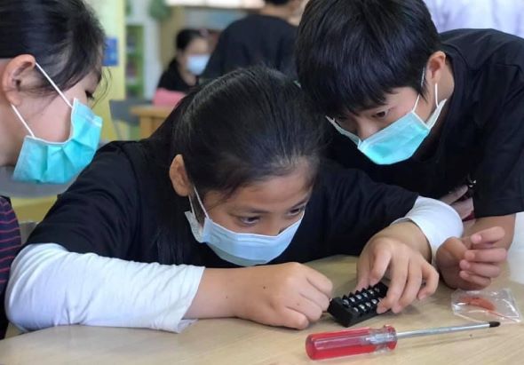 三位戴著口罩和眼鏡的女性在課堂上，正指導一名年輕女學生使用電子元件進行科學實驗。桌上擺放著螺絲起子等工具，展現了教師與學生互動的STEM教育場景。