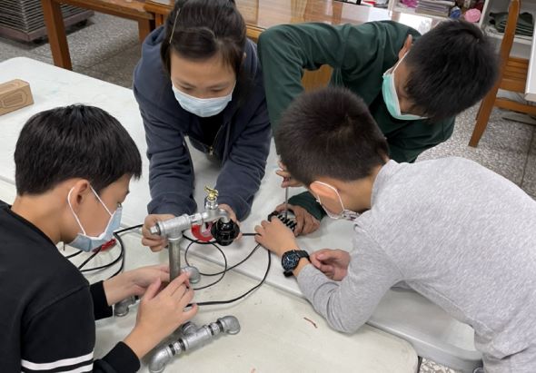 五位戴著口罩的學生圍坐在桌前，一起操作電子機器人和電路板進行實驗。他們專注地組裝和測試電子元件，進行科學教育活動。