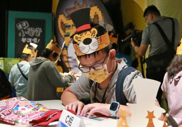 一名戴著老虎頭套和眼鏡的男子坐在桌前簽名或參與活動。桌上放有紅色物品和其他道具。背景有黃色牆面、綠色標牌和其他參與者。這是一場室內主題活動或角色扮演活動的現場。