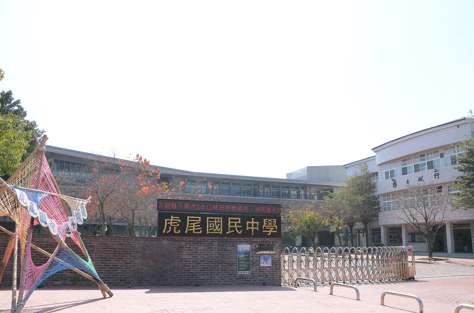 虎尾國民中學校園廣場，紅磚建築物上掛著金色校名牌匾，廣場上有彩色風箏裝飾和伸縮門，右側為白色校舍建築，樹木點綴其間，校園內擺設有紅燈籠，呈現節慶氛圍。