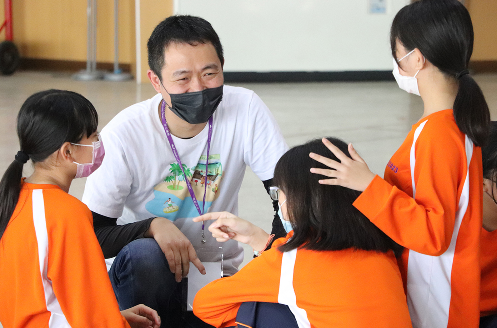 一名戴著黑色口罩、穿著白色上衣的成人教師，蹲坐著與四名穿著橙色運動服的學生互動。教師配戴識別證，正在進行教學或指導活動。背景為室內場景，牆面為米黃色。
