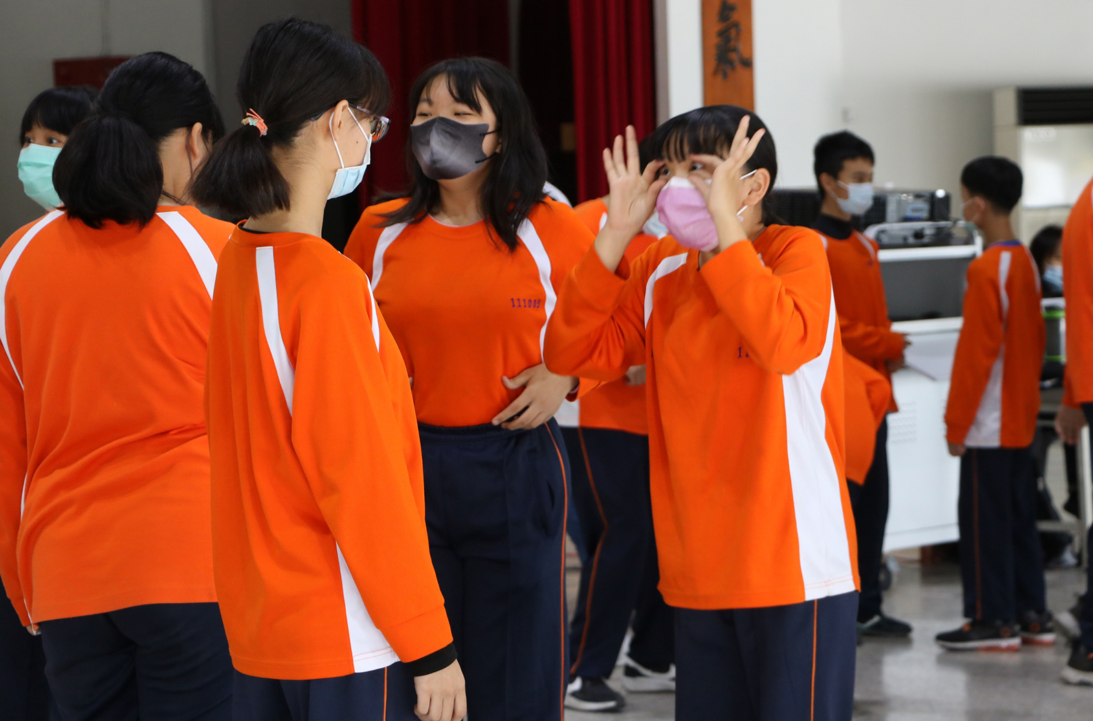 一群學生穿著橙色運動服在室內聚集，多數人戴著口罩。其中一名學生舉起粉紅色物品，做出揮手動作。背景可見紅色布幕和教室設施，其他學生站在一旁觀看。