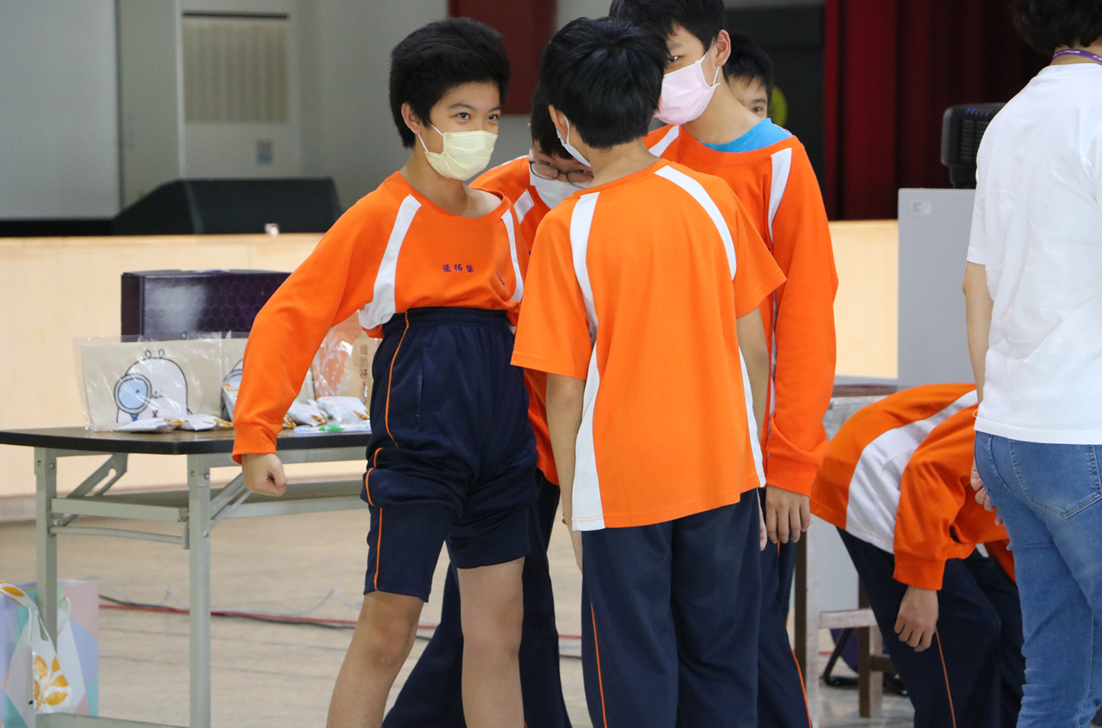 室內體育館中，數名穿著橙色和深藍色運動服的學生戴著口罩聚集一起，似乎在參加學校運動會或體育活動。桌子上放著物品和飲水杯，背景可見其他穿白色衣服的人員。