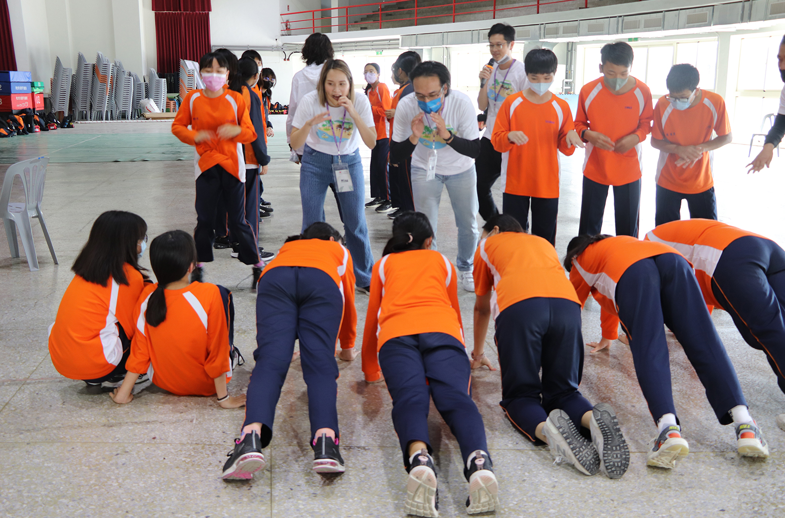 大型室內運動館內，多名穿著橙色和藍色運動服的學生進行俯卧撑訓練。前排學生做俯卧撑，後排學生站立觀看和鼓勵。許多學生戴著口罩。場景展現集體運動訓練和團隊合作的氛圍。