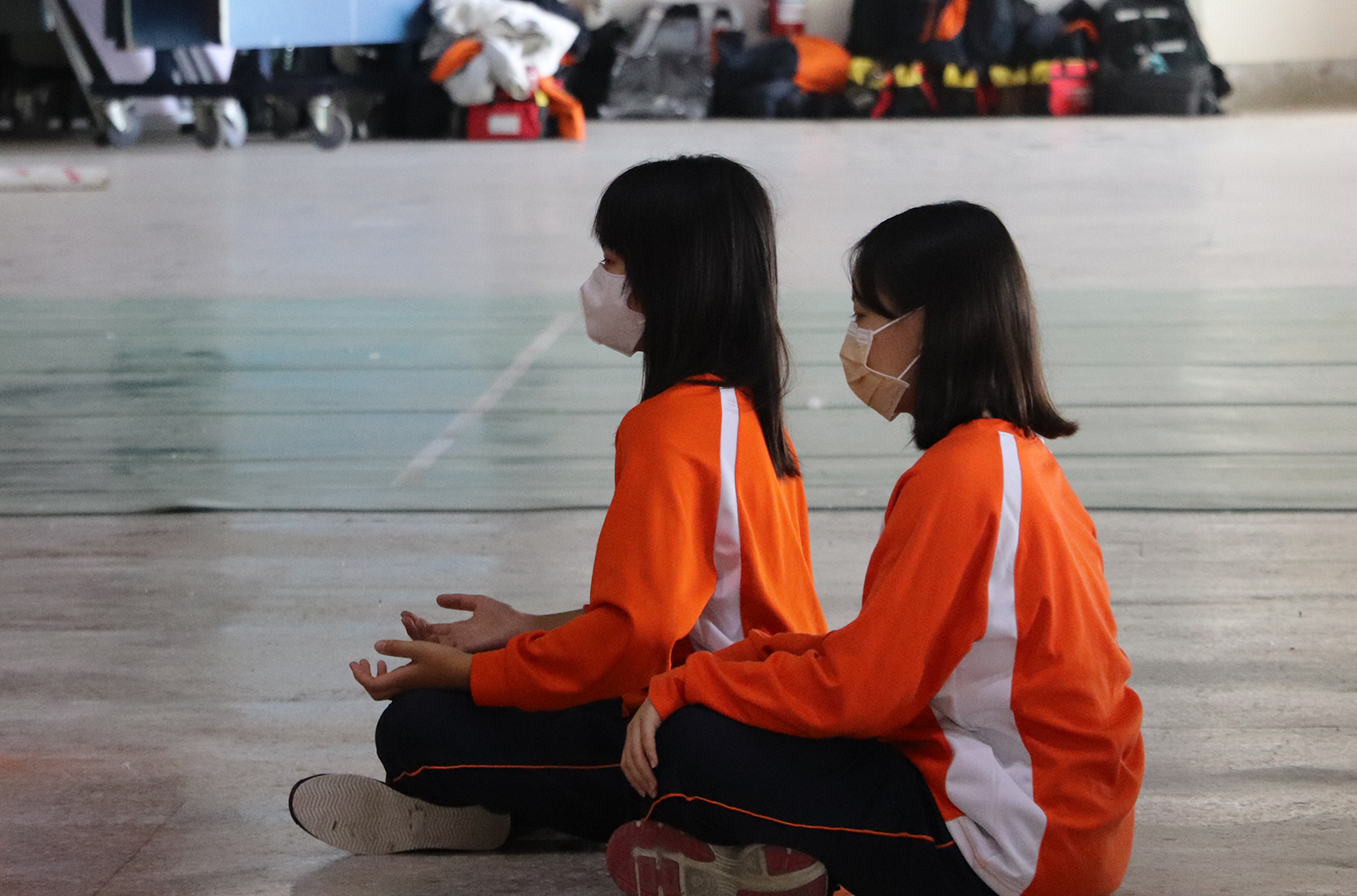 兩名女性身穿橙白色運動服坐在室內場地上，其中一人戴著口罩。背景可見運動器材和裝備。兩人正面向相同方向交談，似乎在進行體育活動或訓練。