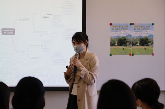 一位戴著口罩的女性講師站在教室前方進行演講，身穿米色外套，雙手做出講解手勢。身後白板上顯示流程圖，右側牆上貼著兩張運動場景的海報。觀眾坐在前排聆聽。