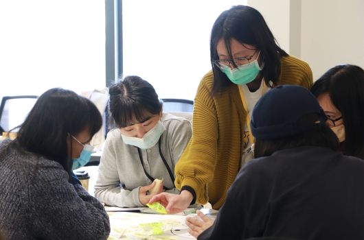 五位戴著口罩的女性在室內聚集在一張桌子周圍，共同查看和討論桌上的文件或資料。她們穿著冬季服裝，認真地進行小組討論或工作會議。背景可見電腦螢幕和窗戶。