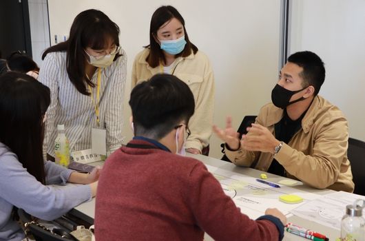室內工作坊現場，五位戴口罩的人員聚集在桌前討論，桌上放有文件、便利貼和筆。左側人員穿著條紋衣，右側人員穿著卡其色衣服，中央穿著紅色毛衣的人坐著傾聽。這是一場團隊協作或培訓活動。