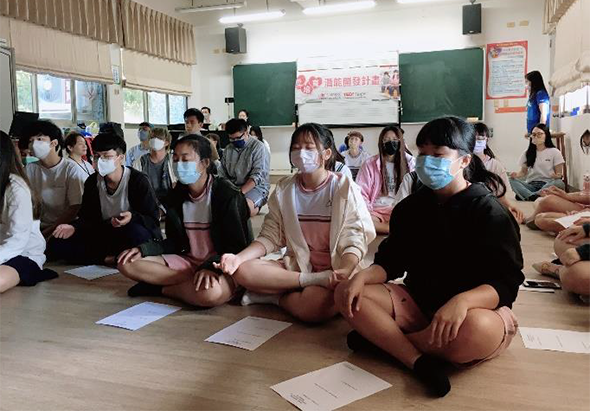 學校教室內，戴著口罩的學生們坐在地板上參加集體活動。多數學生採坐姿面向前方，教室後方有綠色黑板和公告欄，窗戶採光良好，場景呈現師生互動的教學情境。