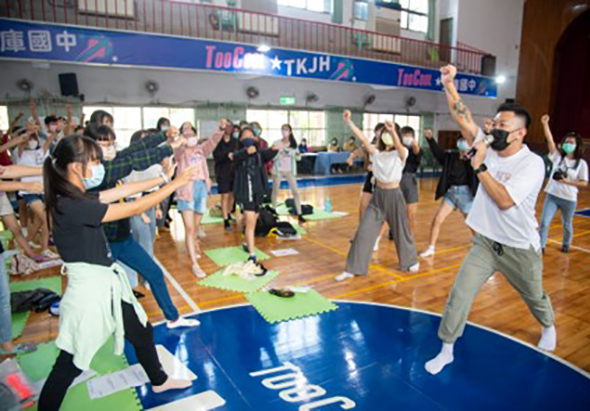 室內體育館中，一群年輕人圍成圓圈進行互動遊戲或團隊建設活動。參與者們舉起雙手，展現熱情參與的姿態。木質地板上鋪有彩色墊子，背景可見藍色的體育館標誌和多位旁觀者。現場充滿活力與歡樂氛圍。