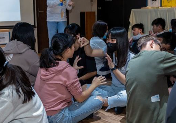 一群年輕人坐在室內地板上圍成一圈，進行團體活動。他們穿著休閒服裝，雙手放在中央相互接觸，展現團隊合作和親密的互動。背景可見投影幕和教室設施，氣氛活躍融洽。