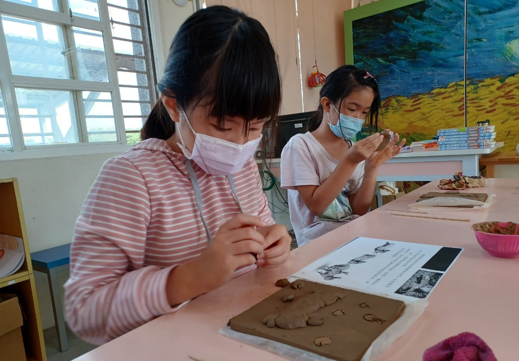 教室內兩名戴口罩的女學生坐在桌前進行陶藝課程。左邊女生正用手指在褐色黏土上刻畫圖案，右邊女生在欣賞和操作黏土。桌上放有教學參考圖片、黏土作品和粉紅色容器。背景有彩繪壁畫和大窗戶。