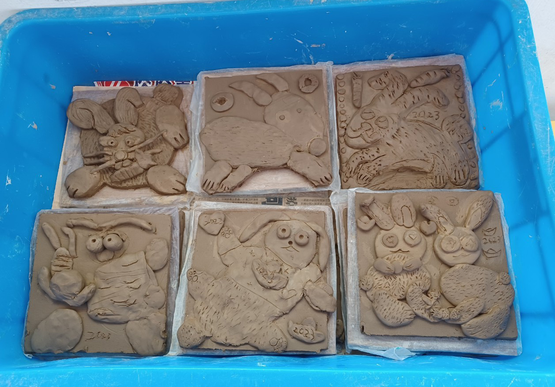 藍色塑膠盒內放置六塊淺褐色黏土雕刻板，上面刻有各式動物浮雕，包括獅子、豬、貓頭鷹、兔子等。每塊板都用白紙包裝保護，板面刻有2023年份標記，展現精細的手工雕刻工藝。