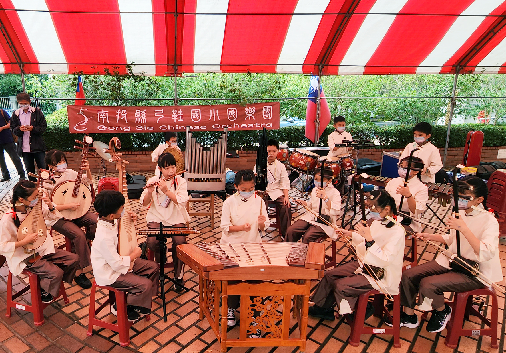 南投縣弓笙樂團在紅白條紋帳篷下進行戶外演奏。團員穿著淺粉紅色制服，演奏琵琶、揚琴、鼓等傳統樂器。舞台背景懸掛台灣國旗與樂團標誌布幕。約二十位學生樂手圍成半圓形座位，正專注演奏傳統國樂。