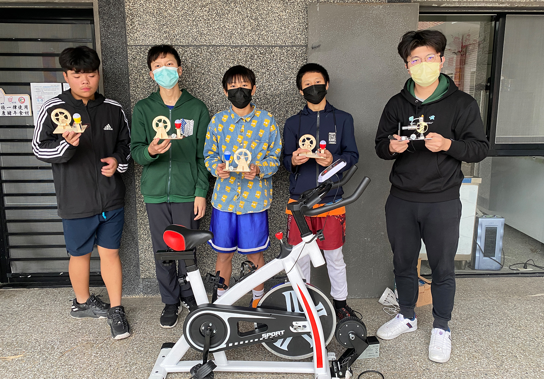 五位年輕人在室內靠牆站成一排，各自手持獎盃。中央放置一台白色健身單車。人們穿著運動服裝，大多戴著口罩。背景是灰色石材牆面和玻璃門窗，地面為淺色瓷磚。這是一場體育競賽或健身活動的得獎合照。