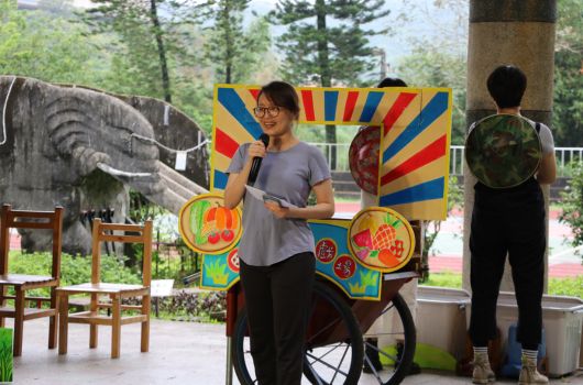 戶外活動現場，一名女性站在彩色風車裝置前展示，背景有大象石雕和樹木，另有參與者背向鏡頭，整體呈現歡樂的兒童遊樂或教育活動氛圍。