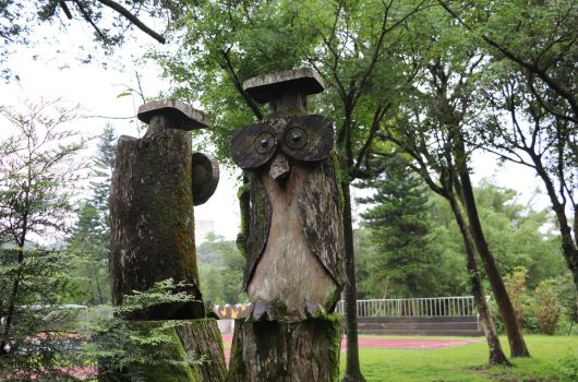 木雕貓頭鷹藝術裝置矗立於公園綠地中，由兩根粗大樹幹雕刻而成，具有圓形大眼睛和尖銳耳朵的特徵。背景為繁茂樹木和遠處的木製欄杆走廊，呈現自然戶外景觀。