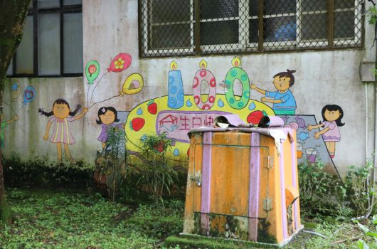老舊建築牆面上的彩色壁畫，描繪手牽手的卡通兒童和彩虹、氣球等圖案。前方放置一個橙黃色條紋的廢棄行李箱，周圍長有雜草。象徵兒童樂園或社區藝術裝置。