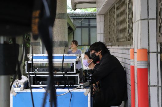 戶外活動現場，兩名戴口罩的工作人員站在藍色桌子後方操作直播設備和麥克風。背景為建築物與玻璃窗，右側可見橘紅色的圓筒物體。前景有黑色設備部分入鏡。