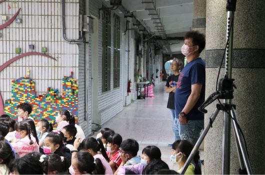幼稚園走廊內，一位老師站在右側為坐在地上的一群幼童講述故事或教學內容。走廊牆上貼有彩色積木裝飾藝術作品。多名幼童穿著粉紅色制服專心聆聽。