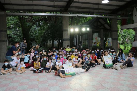 一大群兒童坐在室外廣場上參與活動，舞台上有燈光和背景板，成人協助引導小朋友們，現場熱鬧溫馨，許多小朋友手持材料或作品，呈現集體學習或才藝表演的活動氛圍。