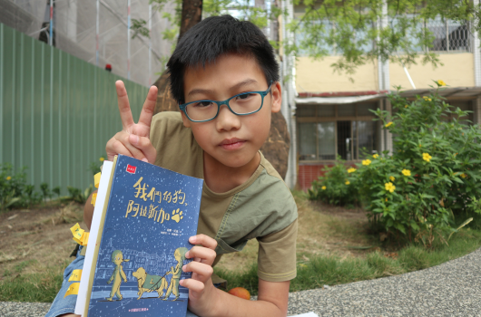 戴著眼鏡的男孩在室外庭園中舉起和平手勢，手持一本藍色封面的書籍，背景是綠色鐵皮牆和開滿黃花的灌木叢。