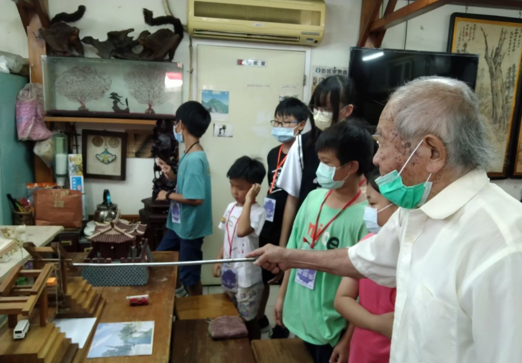 一位年長的男性在傳統文物室內向穿著綠色背心的學生們介紹展品。室內陳列著木雕工藝品、書畫和古董，牆上掛有裝飾品。多名學生戴著口罩聆聽講解，參與文化導覽活動。