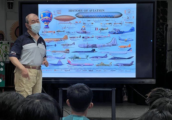 講師戴著口罩站在大螢幕前，展示航空歷史圖表。螢幕上呈現多種飛機機型的演進過程，包括熱氣球、飛船及各時代戰機與民航機。觀眾坐在前方認真聽講。