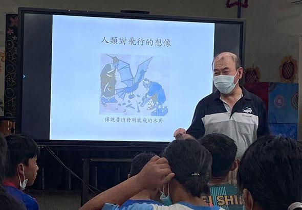 教室內,一位戴著口罩的男性講師站在投影螢幕前向學生講課。螢幕上顯示中文標題「人類對能行的想像」及相關插圖。前景可見多位學生從背後觀看。講師手持教材進行環境教育講座。