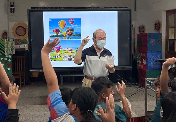 教室內，一位戴著口罩的男性講師在大螢幕前進行教學，螢幕上顯示熱氣球節慶的彩色圖片。眾多學生舉起雙手踴躍參與課堂互動，呈現生動的教學現場。