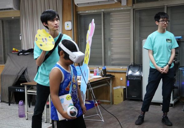 三名學生在教室內進行虛擬實境遊戲體驗。中央學生戴著VR頭盔，揮動控制器；左方學生持遊戲搖桿；右方學生在旁觀看。背景有木製儲物櫃和揚聲器，呈現教育科技應用場景。