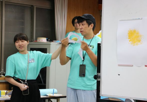 室內教室環境中，兩名穿著青綠色T恤的人員進行教學活動。右側男性講師戴著黑色棒球帽和識別証，舉起藍色和黃色的教具向左側女性示範。牆上貼著黃色圖案，白板和教學用具擺放在室內。