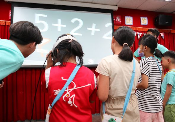 教室內，一群小學生圍在投影幕前，投影幕上顯示數學算式「5+2+?」。學生們穿著不同顏色的衣服，認真地看著題目。紅色窗簾和教室布置可見於背景。