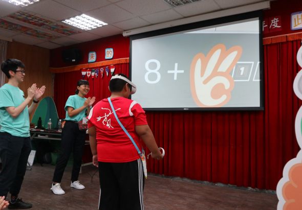 室內教室進行互動數學教學活動，講師在投影幕上展示「8+6=14」的數學題目，搭配手勢圖示。穿著綠色和紅色制服的學生們圍成一圈，積極參與課堂互動。教室牆面貼有紅布裝飾，營造歡樂學習氛圍。