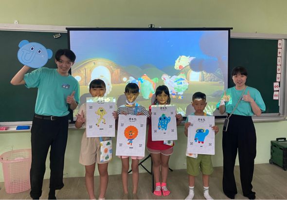 教室內，兩位穿著青色制服的教師與五位小學生展示自製的藝術作品。背景大螢幕播放卡通影片，黑板後方可見教室環境。學生們手持繪有不同圖案的白色紙張作品，呈現創意美術課成果。