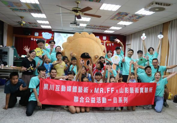 一群穿著青綠色上衣的大人和小孩在室內聚集，共同舉著一面紅色橫幅，橫幅上標示黑川互動媒體藝術、MR.FF與山藝藝術實驗所的聯合公益活動。背景有大型黃色布娃娃裝置和彩色布置。