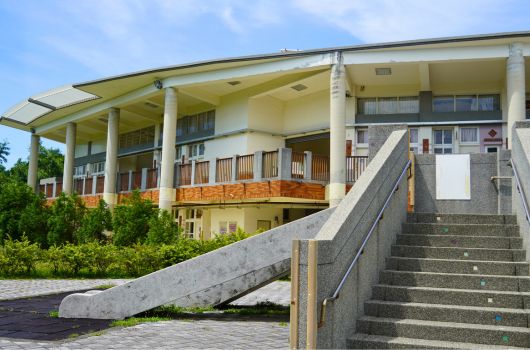 現代建築風格的公共設施，黃色屋頂搭配白色柱子，前方有寬闊的混凝土樓梯和斜坡通道，周圍綠樹環繞，設有無障礙出入口。