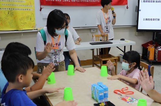 教室內進行教學活動，一位戴口罩的女性教師在綠色杯子旁向坐著的兒童進行互動式教學示範，背景有黃色海報和紅色橫幅，另有兩位教師在旁協助，桌上放有教具和玩具。