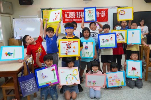 幼兒園教室內，約二十位小朋友展示自己繪製的彩色畫作。孩子們穿著紅、藍、黃等鮮豔服裝，手持或站在自己的畫框前。牆上貼有中文標語和裝飾品。地板上也放置多幅兒童畫作，呈現溫馨的藝術展示活動氛圍。
