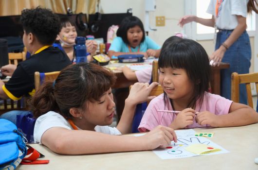 教室內兩名女學生坐在桌前，一起觀看和討論繪畫作品。背景可見其他學生和成人在進行活動。前景學生穿著藍色和粉紅色衣服，專注地看著桌上的圖畫，展現友善的互動和學習氛圍。