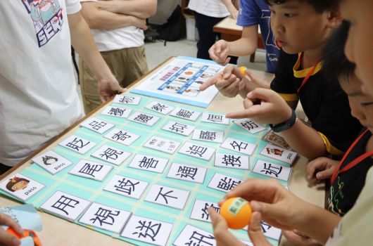 一群學生圍坐在桌前，一起進行漢字學習遊戲。桌上鋪著綠色遊戲板，上面排列著多個漢字卡片，學生正用手指指著卡片互動學習。背景有其他學生和老師站立觀看，呈現生動的課堂教學活動場景。