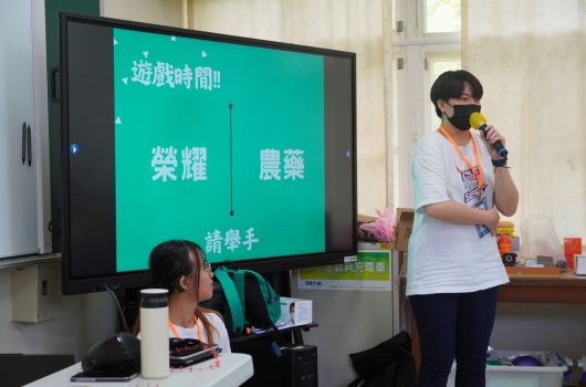 教室內，一位戴口罩的女性講者站在大螢幕前進行簡報。螢幕顯示綠色背景的中文投影片，標題為「遊戲時間」，內容涵蓋「榮耀」、「農藥」等主題。講桌前坐著一位學生聽眾。