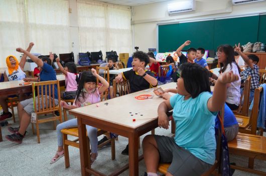 一間教室內，多名小學生圍坐在木製課桌旁，舉起雙手參與互動式課堂活動。教室後方放置電腦，綠色黑板掛在牆上，學生們看起來正積極地參與課程或遊戲。